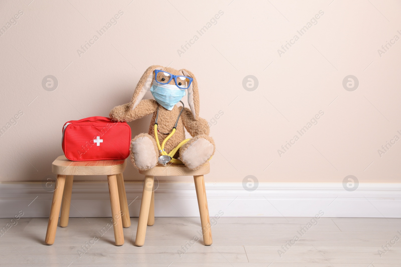 Photo of Toy bunny with eyeglasses, stethoscope and first aid bag near beige wall, space for text. Pediatrician practice
