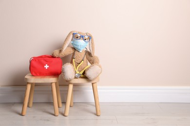 Toy bunny with eyeglasses, stethoscope and first aid bag near beige wall, space for text. Pediatrician practice