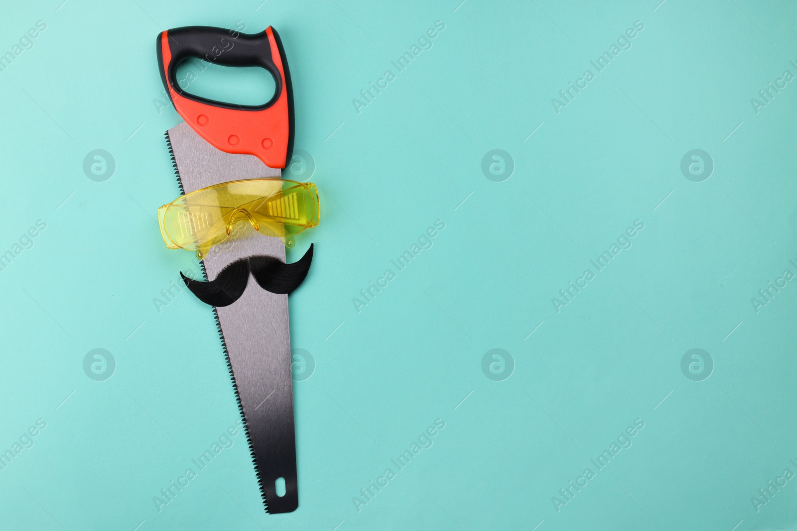 Photo of Man's face made of artificial mustache, safety glasses and hand saw on light blue background, top view. Space for text