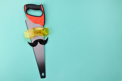 Photo of Man's face made of artificial mustache, safety glasses and hand saw on light blue background, top view. Space for text