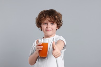 Cute little boy with glass of fresh juice showing thumb up on light gray background