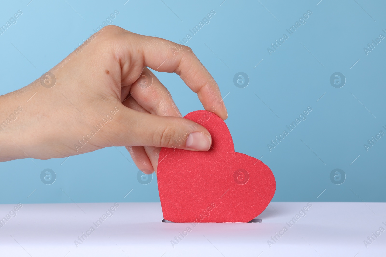 Photo of Woman putting red heart into slot of donation box against light blue background, closeup
