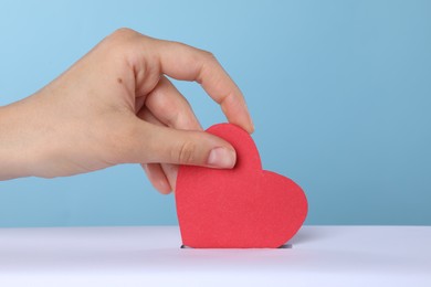 Woman putting red heart into slot of donation box against light blue background, closeup