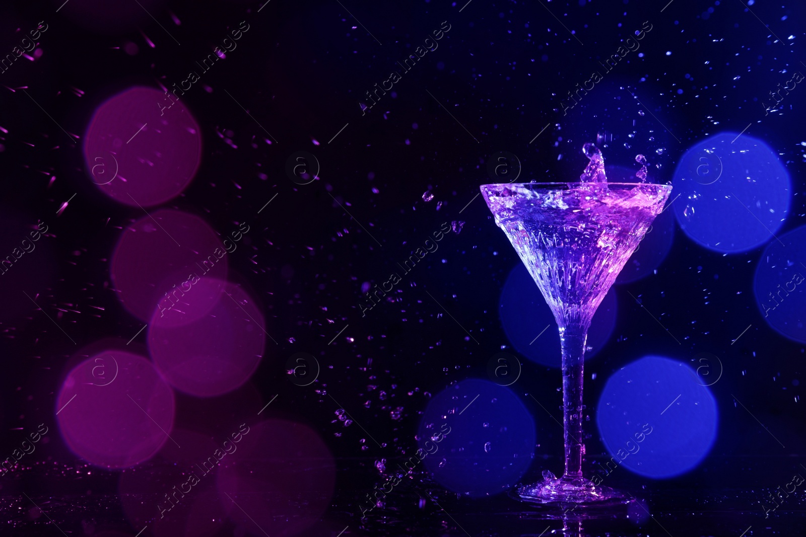 Photo of Glass of martini and splashes in neon lights on dark background, bokeh effect with space for text