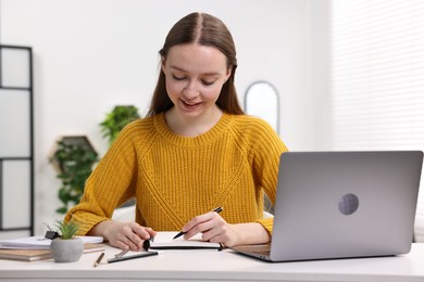 Photo of E-learning. Young woman taking notes during online lesson at white table indoors