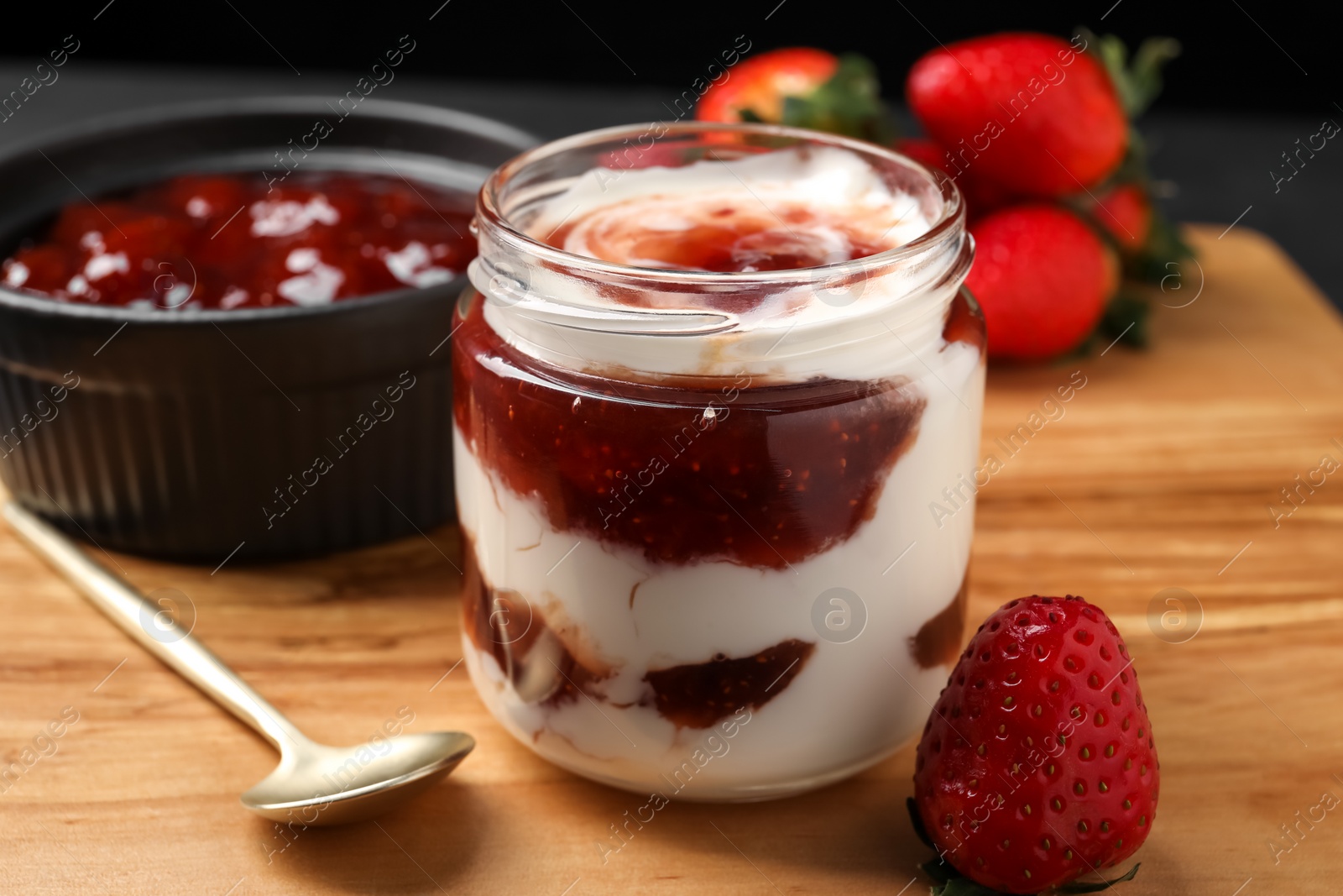 Photo of Tasty yoghurt with jam, spoon and strawberries on wooden board, closeup