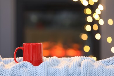 Photo of Cup of hot drink on knitted blanket near fireplace at home, space for text. Cozy atmosphere