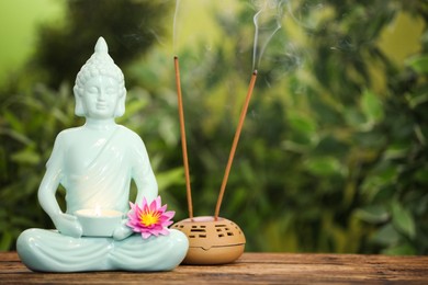 Photo of Buddha statue with burning candle and lotus flower near incense sticks on wooden table. Space for text