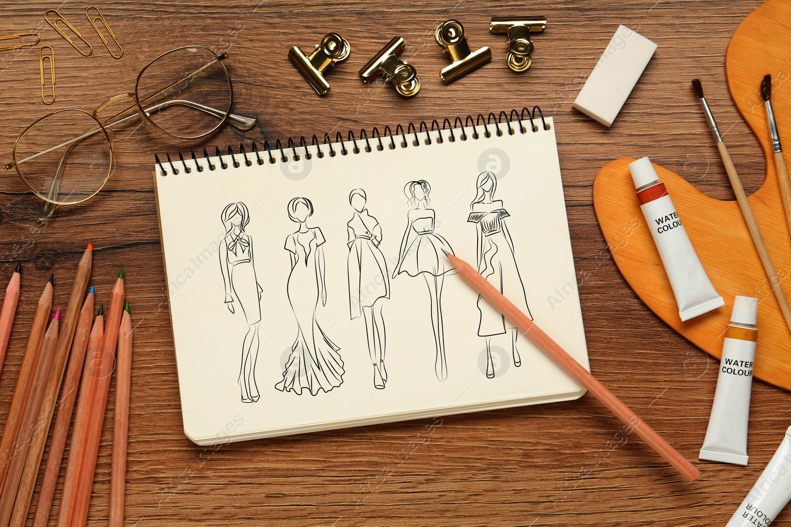 Image of Sketches of different clothes in pad on wooden table. Fashion designer's desk with stationery, flat lay