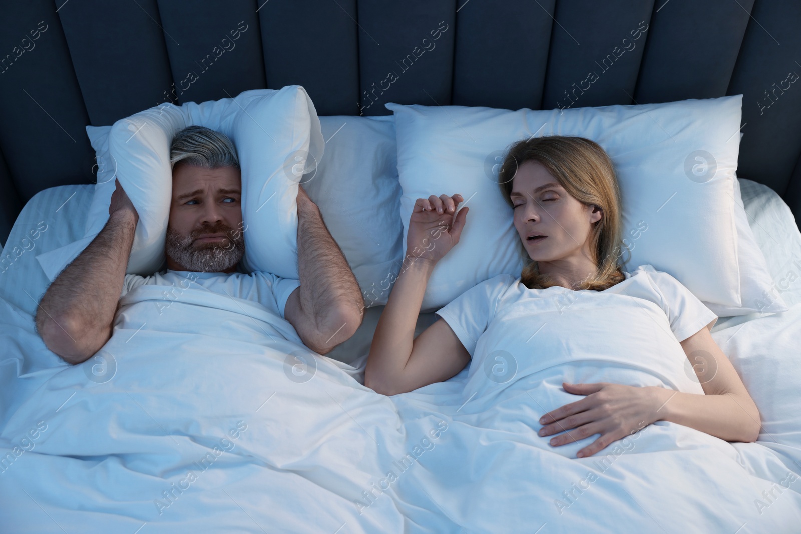Photo of Irritated man covering his ears with pillows in bed at home. Problem with snoring wife
