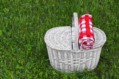 Photo of Rolled checkered tablecloth with picnic basket on green grass outdoors, space for text