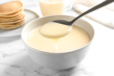 Photo of Spoon of pouring condensed milk over bowl on marble table, closeup. Dairy products