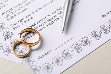 Marriage contract, gold rings and pen on light wooden table, closeup