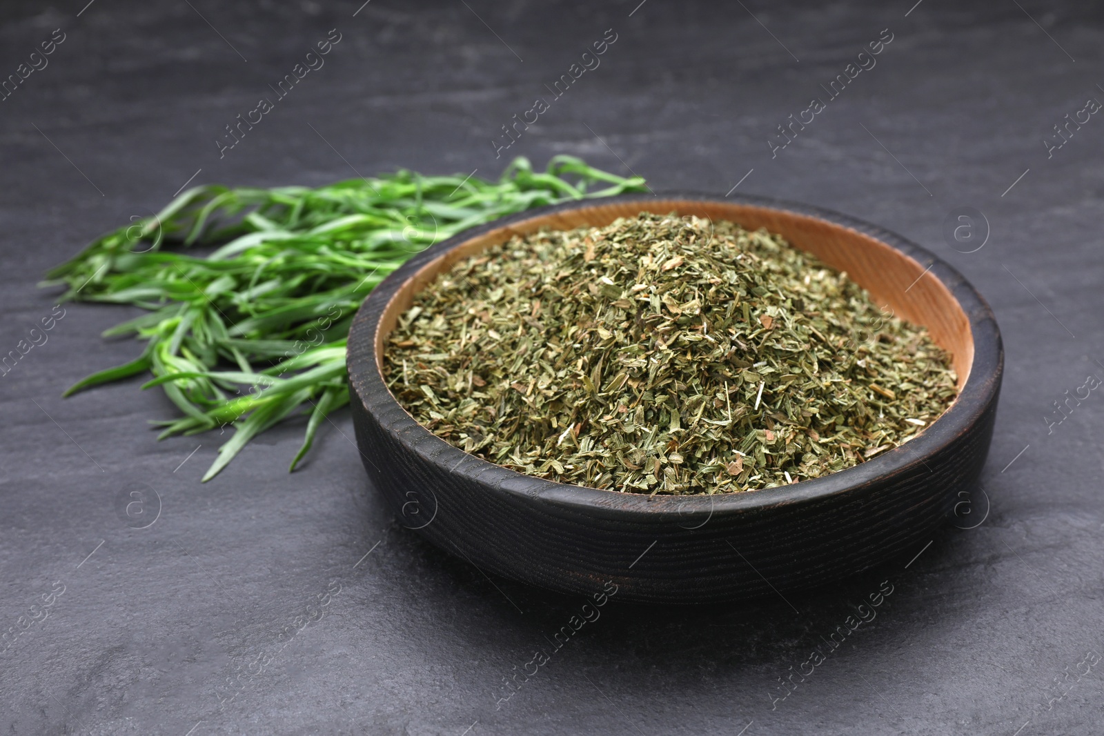 Photo of Dry tarragon in bowl and green leaves on black textured table, closeup