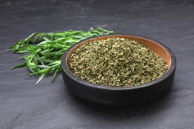 Photo of Dry tarragon in bowl and green leaves on black textured table, closeup