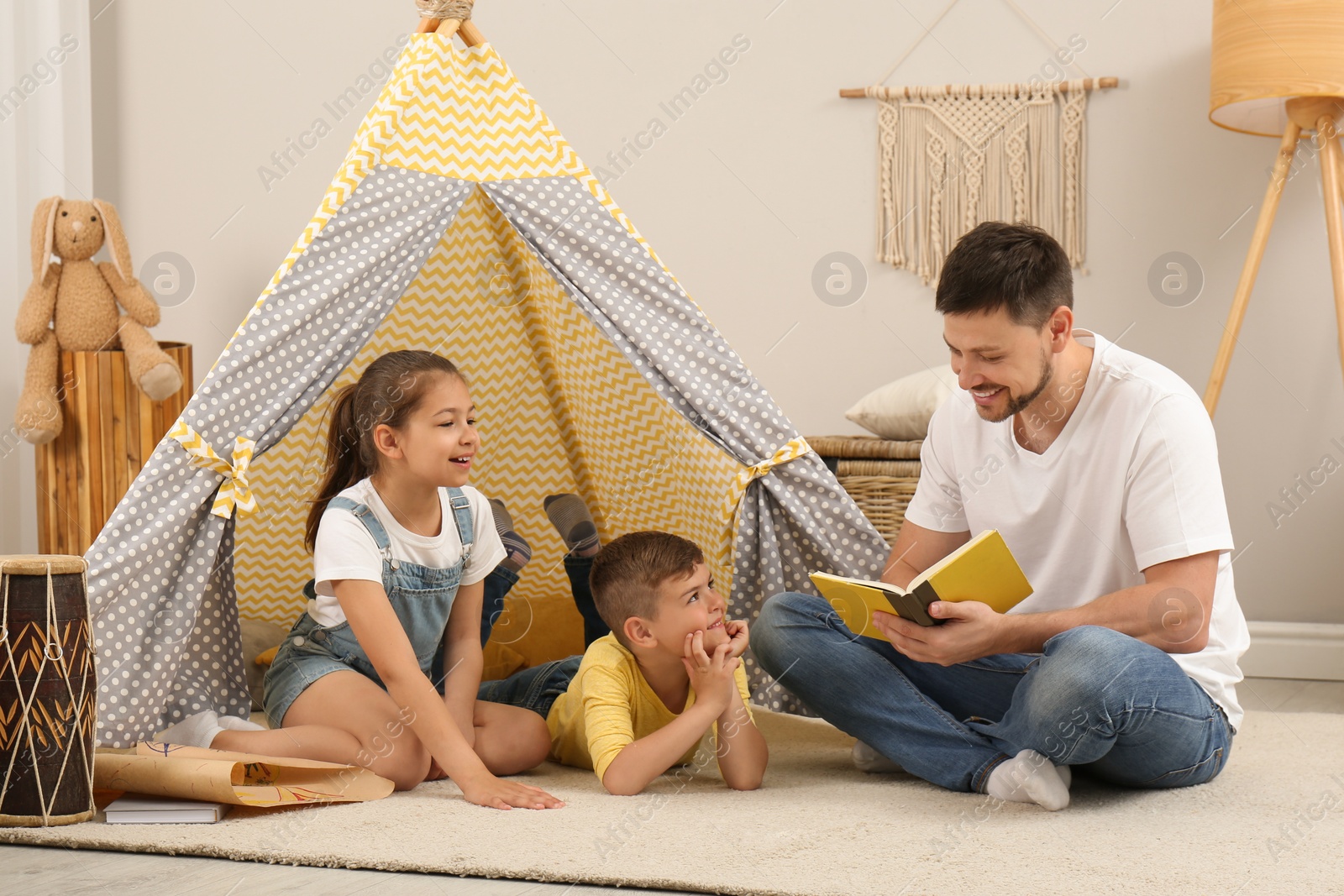 Photo of Father reading book to children near toy wigwam at home