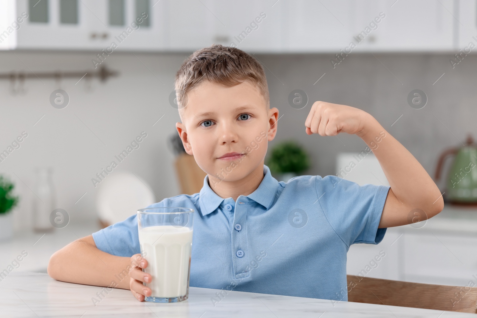 Photo of Cute boy with glass of fresh milk showing his strength at white table in kitchen