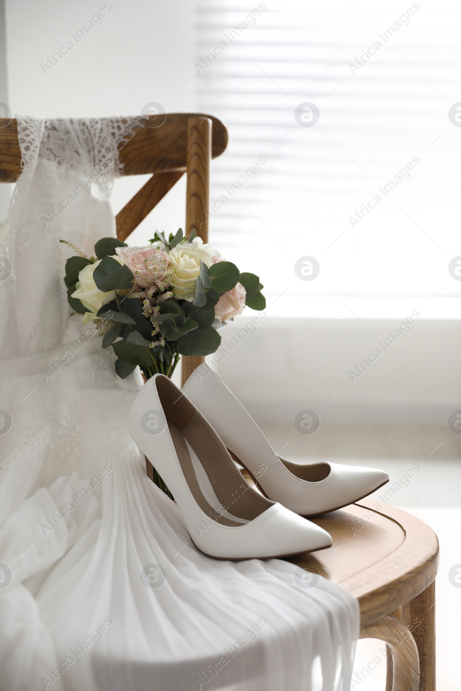 Photo of White high heel shoes, flowers and wedding dress on wooden chair indoors