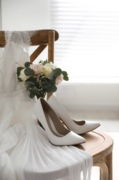 Photo of White high heel shoes, flowers and wedding dress on wooden chair indoors
