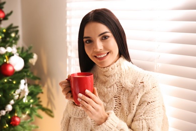 Photo of Young woman holding cup of coffee in room with Christmas tree
