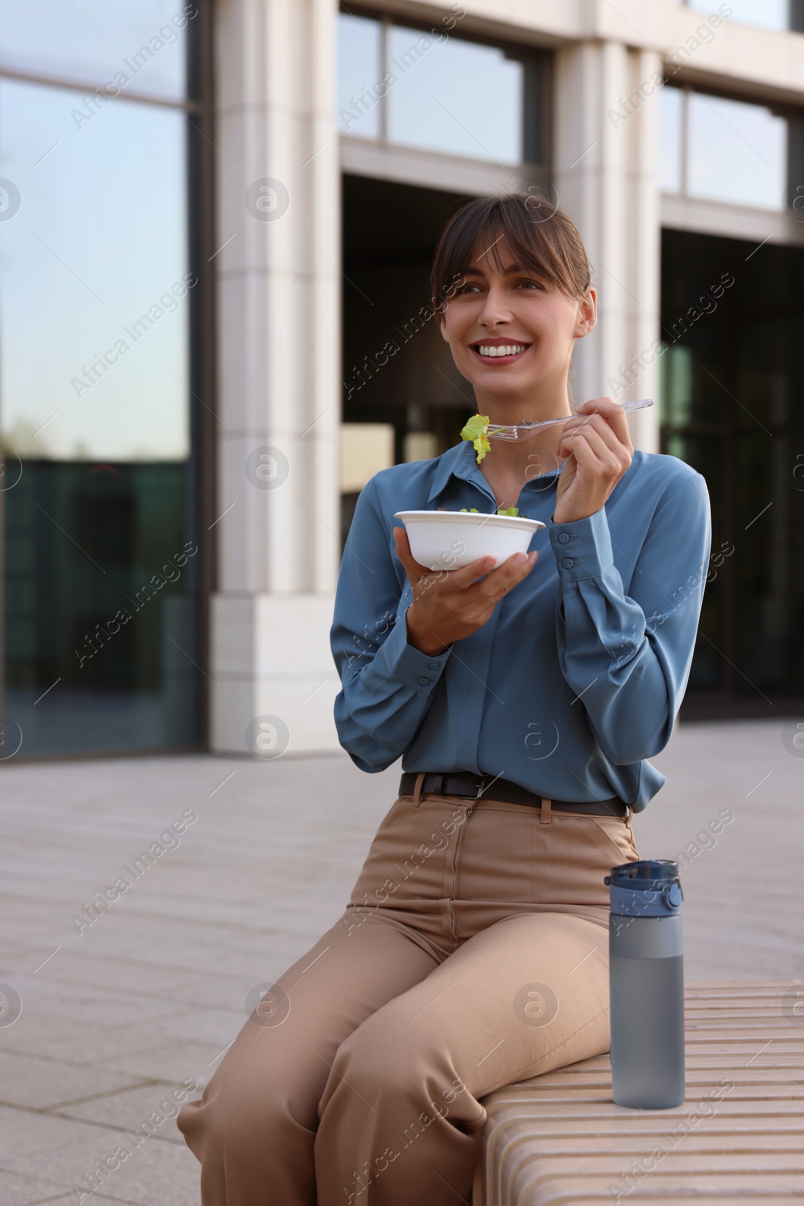 Photo of Happy businesswoman with plastic bowl of salad and bottle of water having lunch on bench outdoors