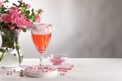 Cotton candy cocktail in glass, marshmallows, vase with pink roses and straws on white wooden table against gray background, space for text