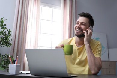 Photo of Young man talking on phone while working with laptop at desk in home office