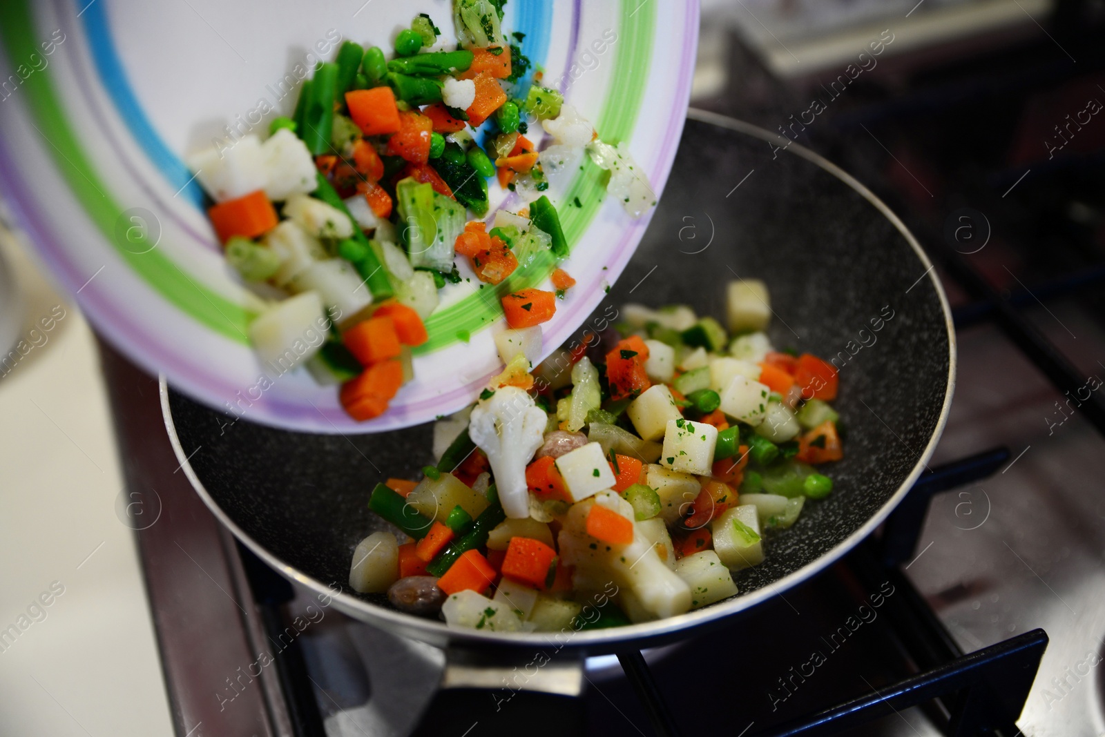 Photo of Pouring mix of fresh vegetables into frying pan on kitchen stove, closeup