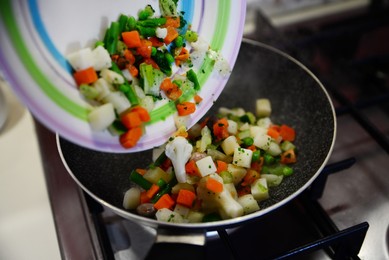 Pouring mix of fresh vegetables into frying pan on kitchen stove, closeup