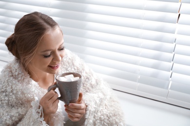 Photo of Beautiful young woman with cup of hot drink and marshmallow near window at home. Winter atmosphere