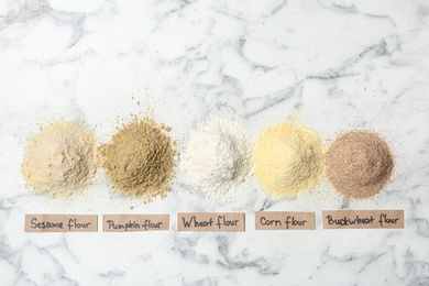Piles of different flour types with tags on marble background, top view