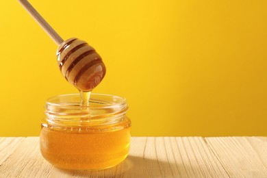 Pouring honey from dipper into jar at wooden table against yellow background, space for text