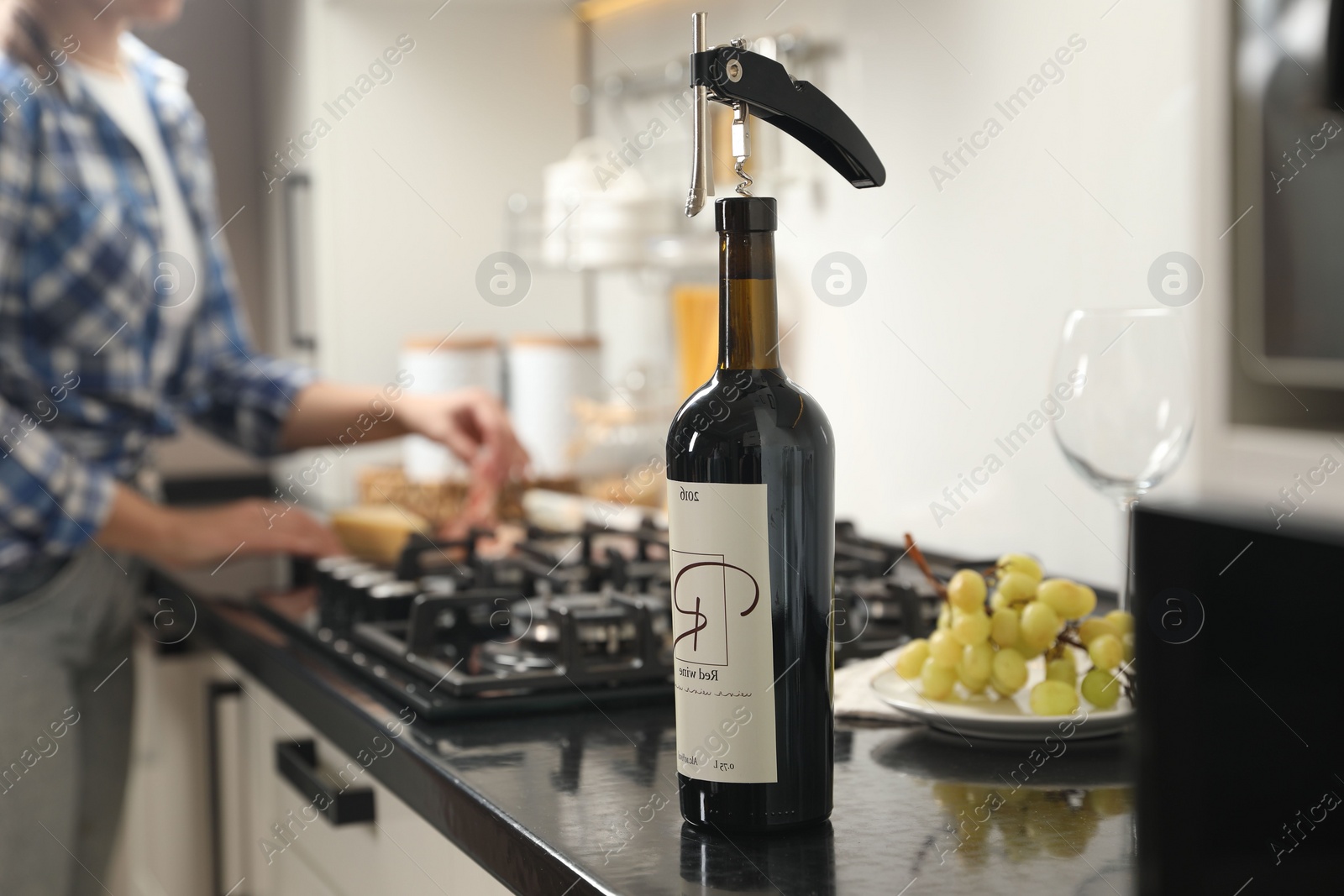 Photo of Wine bottle with corkscrew on black countertop, closeup. Woman in kitchen, selective focus