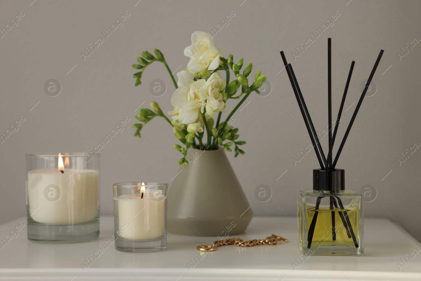 Photo of Aromatic reed air freshener, freesia flowers and candles on white bedside table indoors