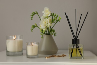 Photo of Aromatic reed air freshener, freesia flowers and candles on white bedside table indoors