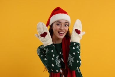 Photo of Happy young woman in Christmas sweater, Santa hat and knitted mittens on orange background