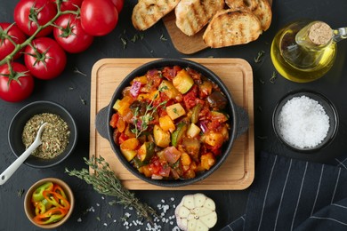 Photo of Dish with tasty ratatouille, ingredients and bread on black table, flat lay