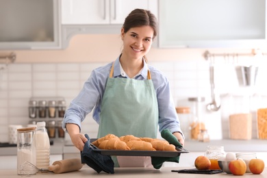 Woman holding baking tray with delicious croissants in kitchen