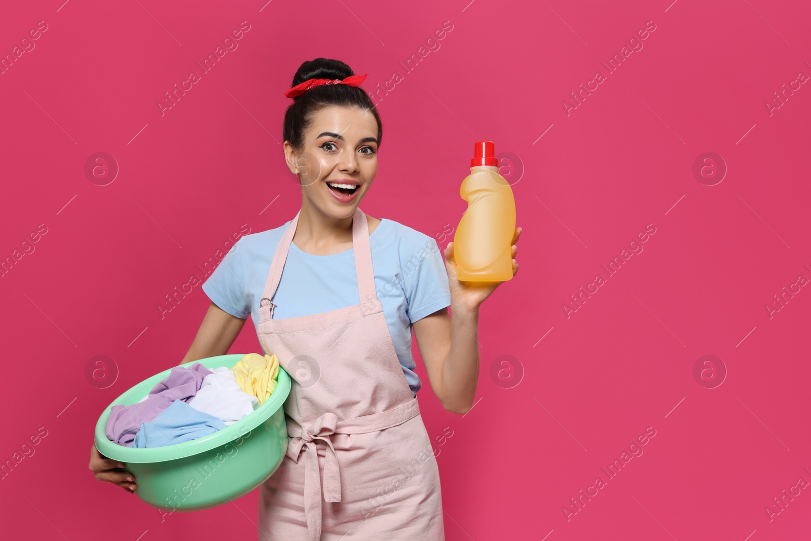 Photo of Housewife holding bottle of cleaning product and basin with clothes on pink background, space for text