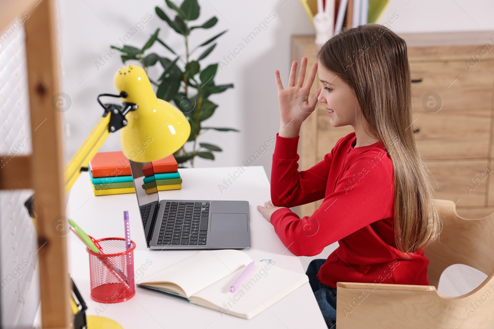 Photo of E-learning. Cute girl raising her hand to answer during online lesson at table indoors