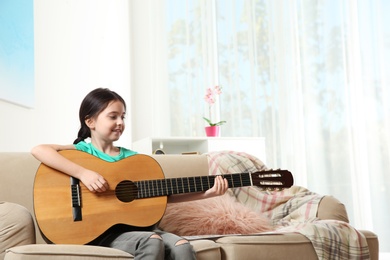 Cute little girl playing guitar on sofa in room. Space for text