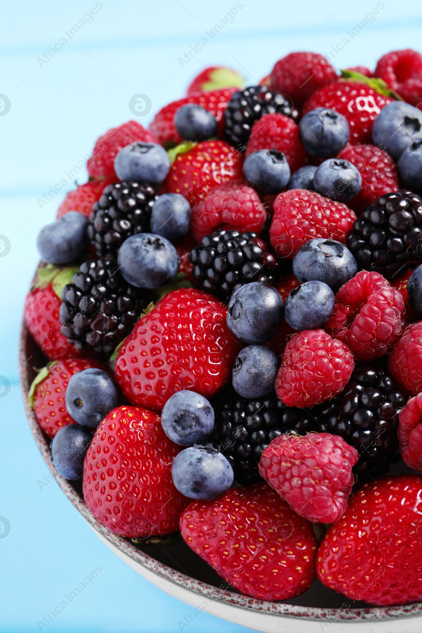 Photo of Different fresh ripe berries in bowl on light blue table, closeup