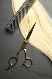 Photo of Professional hairdresser scissors and comb with blonde hair strand on dark grey table, flat lay