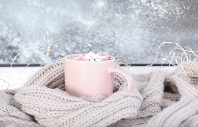 Cup of aromatic cacao with marshmallows and   warm scarf on window sill