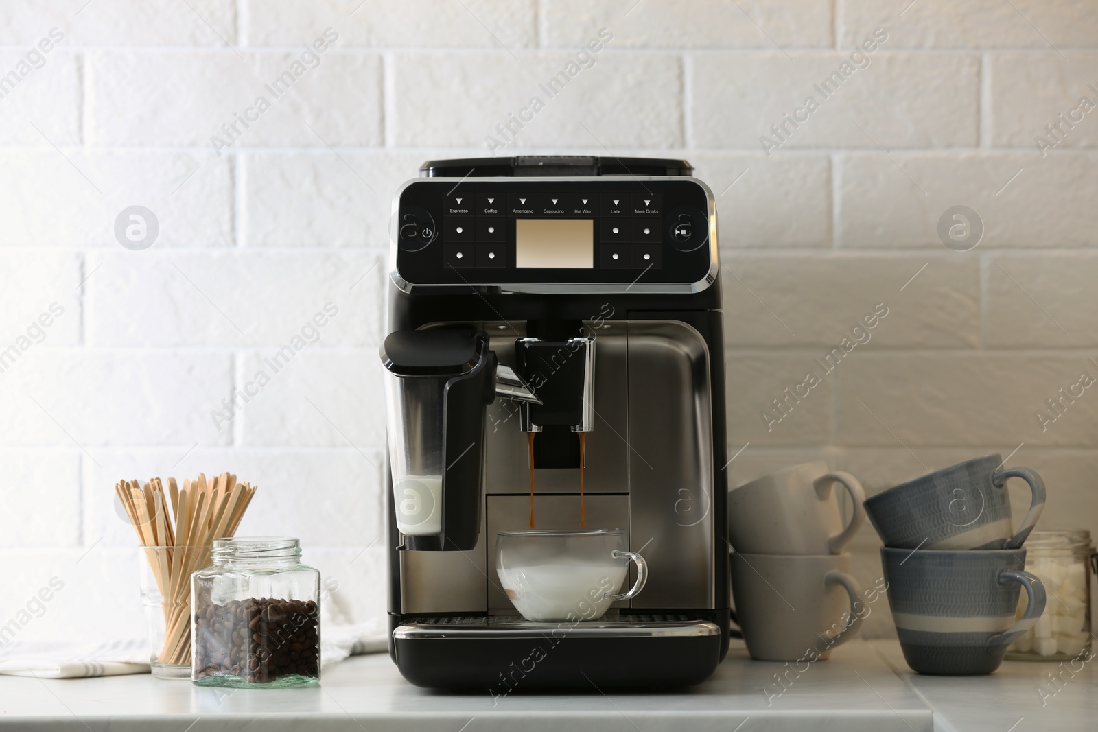 Photo of Modern espresso machine pouring coffee into glass cup with milk on white countertop in kitchen