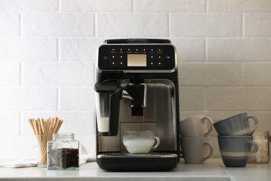Photo of Modern espresso machine pouring coffee into glass cup with milk on white countertop in kitchen