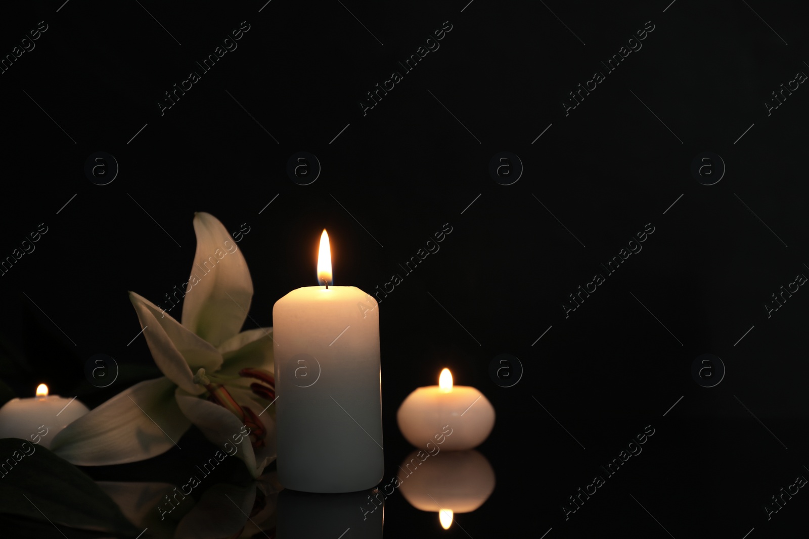 Photo of White lily and burning candles on black mirror surface in darkness, space for text. Funeral symbols
