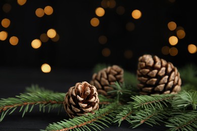 Photo of Christmas greeting card with space for text. Fir tree branches and pine cones on wooden table against blurred festive lights