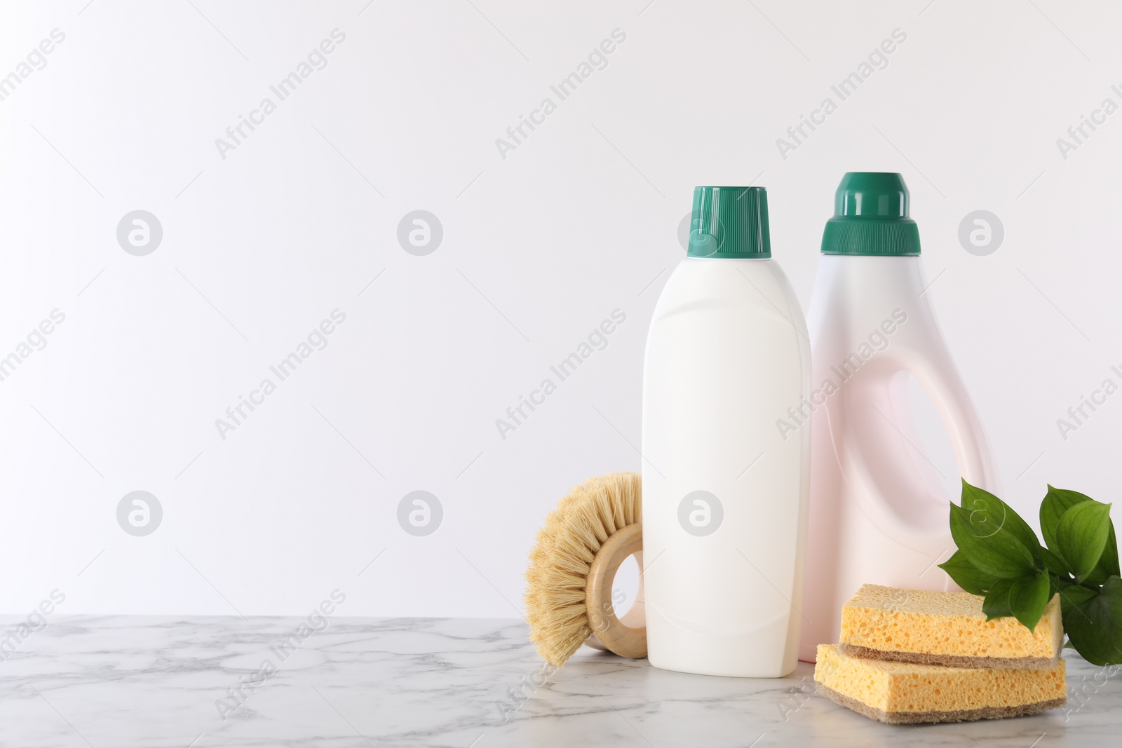 Photo of Bottles of cleaning product, brush, sponges and green leaves on white marble table. Space for text