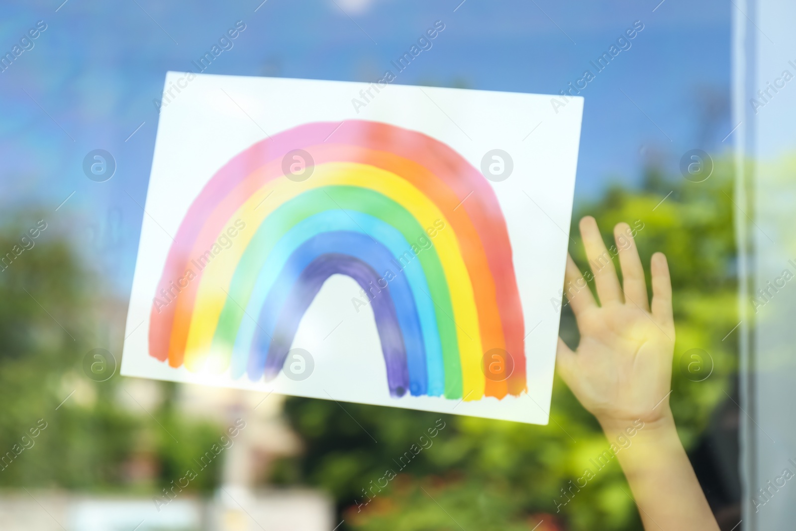 Photo of Closeup of little girl with rainbow picture near window, view from outdoors. Stay at home concept